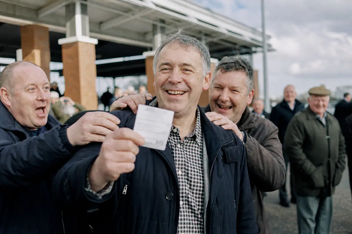 Un homme souriant tient un ticket gagnant devant un hippodrome